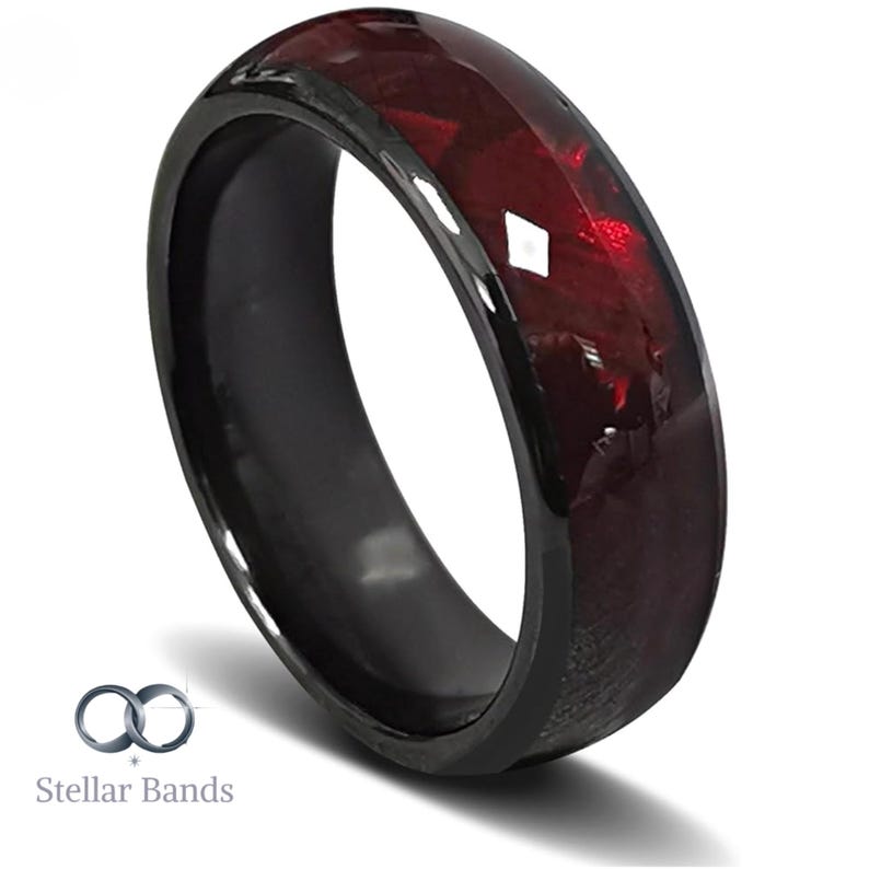 Puede incluir: Un anillo negro con un centro facetado de color rojo intenso. El anillo tiene un acabado pulido y brillante. El nombre de la marca "Stellar Bands" es visible en la esquina inferior izquierda.