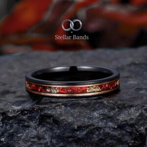 May include: A black ring with a red and gold inlay. The ring has a polished black band with a thin gold-colored border. The inlay features a vibrant red stone with gold flecks. The ring is from Stellar Bands.