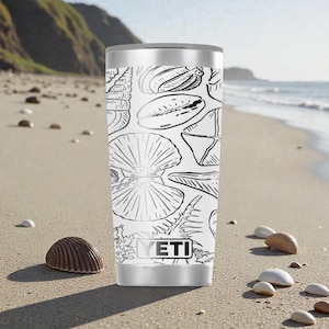 Puede incluir: Un vaso térmico blanco y plateado con un dibujo lineal negro de varias conchas marinas. El vaso tiene el nombre de la marca "YETI" impreso en la parte delantera. El fondo muestra una playa arenosa con conchas.