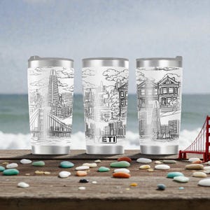 Puede incluir: Tres vasos de viaje de acero inoxidable con dibujos lineales en blanco y negro de los monumentos de San Francisco. Se muestran el Golden Gate Bridge, la Pirámide Transamerica y casas victorianas. Los vasos tienen tapas y bases plateadas, y la marca "YETI" es visible.