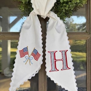 May include: White fabric door hanger with scalloped edges, embroidered with crossed American flags and the letter "H" in red and blue. The hanger is tied to a green wreath, creating a patriotic decorative piece.