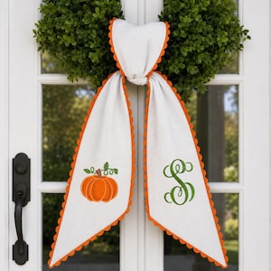May include: A white fabric door hanger featuring a scalloped orange trim. The left side displays an embroidered orange pumpkin with green leaves and vines, while the right side features a large, decorative green embroidered letter S monogram.