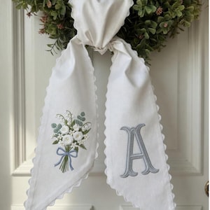 Pode incluir: Cabide de porta de tecido branco com borda ondulada. Um lado apresenta um buquê de rosas brancas e bordado de fita azul. O outro lado tem uma letra "A" cinza bordada.