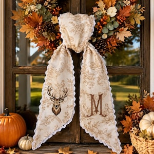 May include: A cream-colored fabric scarf featuring a brown toile de jouy pattern, a stag head illustration on one side, and a large letter M on the other. The scarf has a white scalloped lace trim along the edges and is draped over a window.