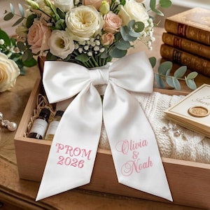Könnte beinhalten: Ein weißes Satinband mit rosa gesticktem Text "PROM 2026" und "Olivia & Noah". Das Band ist Teil einer Geschenkbox-Anordnung mit einem Rosenstrauß, einem weißen Pullover und kleinen Flaschen.