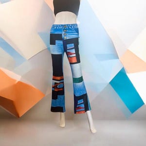 May include: High-waisted flared pants featuring a blue denim print with a button fly closure. The fabric displays a patchwork design of black, blue, orange, and green geometric shapes and abstract blocks, creating a bold, multi-colored visual pattern.