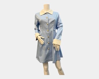 Puede incluir: Un abrigo vestido azul claro con cuello y puños blancos. El abrigo tiene mangas largas y se abrocha en la parte delantera con botones dorados. El vestido se exhibe en un maniquí.