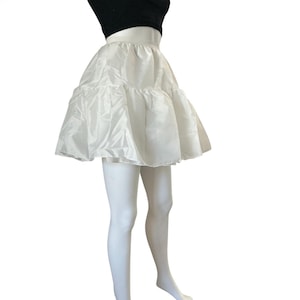 May include: A white petticoat skirt featuring a high-waisted design with a voluminous, tiered silhouette. The garment is crafted from a smooth, slightly shiny fabric that creates a full, flared shape, extending to mid-thigh length with a gathered hem.