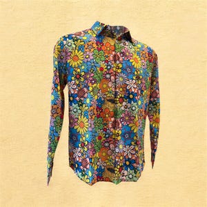 May include: A long-sleeved button-down shirt with a vibrant floral pattern. The shirt features a colorful array of flowers in shades of blue, yellow, orange, pink, and green. The collar and cuffs are visible, and the shirt is set against a light yellow background.