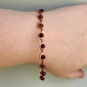 May include: A bracelet featuring a wavy copper wire frame with small, round, faceted dark red beads secured at intervals along the length of the wire. The wire is twisted around each bead to hold them in place, creating a continuous serpentine pattern.