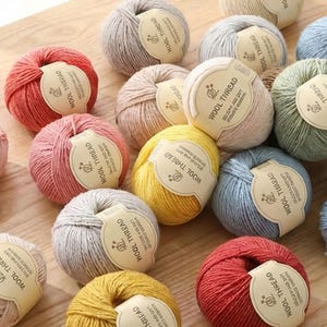 Op de afbeelding: Een verzameling kleurrijke bollen garen in tinten rood, roze, geel, blauw en grijs. Elke bol heeft een papieren label met de tekst "WOOL THREAD". Het garen is gerangschikt op een lichtbruin houten oppervlak.