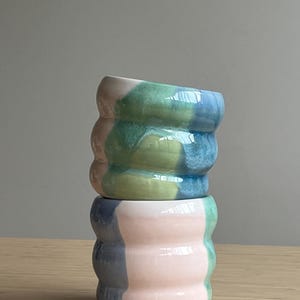 Może przedstawiać: Dwa ustawione jeden na drugim ceramiczne kubki o falistym, żebrowanym kształcie. Górny kubek ma mieszankę zielonych, niebieskich i delikatnie różowych szkliw. Dolny kubek prezentuje wzór z bloków kolorów: jasnego różu, przygaszonego błękitu i zieleni.