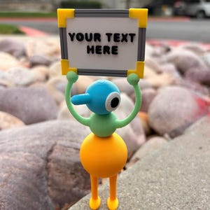 May include: A small, colorful 3D-printed figurine with a round orange base, light green torso and arms, and a blue head with a single eye. The figure holds a rectangular white sign with a gray border and yellow corners, featuring the text YOUR TEXT HERE.