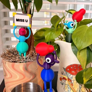 May include: Three colorful, stylized figurines with round bodies and large eyes. One blue and teal figure holds a sign reading MISS U. A purple and blue figure holds a red object. A light green figure holds a red object. The figures are placed among indoor plants.