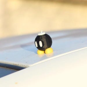 Puede incluir: Figura de bomba de dibujos animados negra con ojos blancos, una mecha blanca y pies amarillos. La figura esférica está sobre un fondo borroso. El objeto es un objeto de colección.