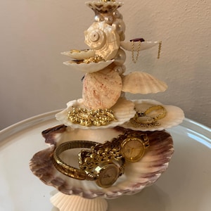 May include: A tiered jewelry organizer constructed from various white and tan seashells and cream-colored pearls. The structure features multiple shell platforms holding gold-toned watches, necklaces, and rings, with a large scalloped shell base.
