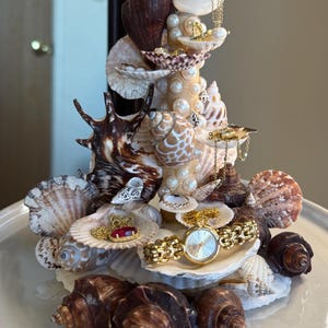 May include: A decorative seashell sculpture featuring various seashells, pearls, and gold jewellery, including a watch and necklaces. The sculpture is arranged on a white plate, showcasing a variety of shell shapes and colours.