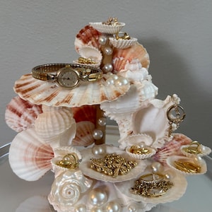 May include: A multi-tiered jewelry organizer constructed from various white and pink-toned seashells. The structure features several shell platforms adorned with white pearls, holding a gold-tone wristwatch, gold chains, rings, and earrings.