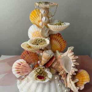 May include: A decorative jewelry stand constructed from various seashells and white pearls. The structure features multiple shell tiers serving as small trays for holding gold chains, rings, and a pendant with a red stone. The base is a large white scallop shell.