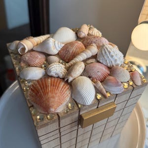 Może przedstawiać: Drewniane pudełko ozdobione różnorodnymi muszlami w odcieniach bieli, różu i beżu. Pudełko posiada złote zapięcie i jest ozdobione małymi, przezroczystymi kamieniami. Pudełko jest przedmiotem dekoracyjnym.