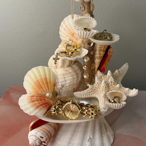 Può includere: Un portagioielli a conchiglia con vari anelli dorati, un orologio e una collana. Il portagioielli è fatto di conchiglie e stelle marine, con una colonna centrale in legno adornata con strass. I gioielli sono esposti su piccole piattaforme di conchiglie bianche.