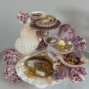 May include: A tiered jewelry organizer constructed from various seashells, including a large central spiral shell and multiple purple-spotted scallop shells. The structure holds gold-toned jewelry, including a watch, chains, and rings, accented with white pearls.