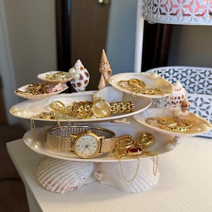 May include: A tiered jewelry organizer made from seashells, displaying gold watches, necklaces, bracelets, and rings. The organizer is adorned with pearls and various seashells, creating a coastal theme. The jewelry is arranged on white shell-shaped trays.