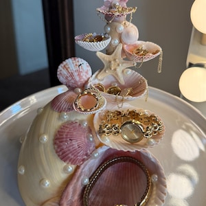 Könnte beinhalten: Ein Muschel-Schmuck-Organizer mit mehreren Ebenen, verziert mit rosa und cremefarbenen Muscheln, Perlen und einem Seestern. Der Organizer enthält Goldschmuck, darunter Ringe, Halsketten und eine Uhr. Die Basis ist ein weißer Teller.