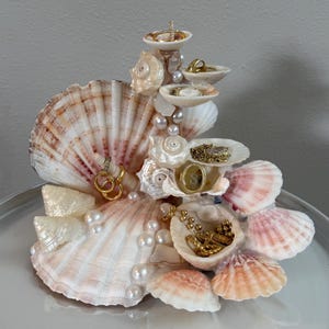 May include: A decorative jewellery holder constructed from various white and pink-toned seashells and scattered white pearls. The structure features multiple shell tiers and protrusions designed to display rings, necklaces, and other small accessories.
