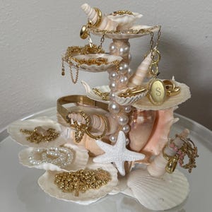 May include: A multi-tiered jewelry organizer constructed from various white seashells and a central column of white pearls. The stand features several shell tiers holding gold chains, gold hoop earrings, a gold oval pendant with the letter S, and pearl bracelets.