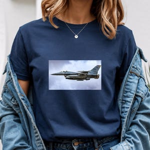 May include: A navy blue short-sleeved t-shirt featuring a rectangular graphic print of a grey fighter jet flying through a cloudy sky. The shirt is worn under a blue denim jacket, and a thin silver necklace with a small circular pendant is visible.