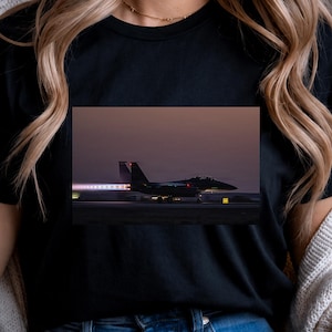 May include: A black t-shirt featuring a graphic print of a fighter jet on a runway at dusk. The jet is shown in profile with its engines ignited, emitting a bright exhaust flame. The scene captures the aircraft during takeoff or taxiing.