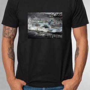 May include: A black short-sleeved t-shirt featuring a rectangular graphic print on the chest. The image depicts two gray fighter jets positioned on the flight deck of an aircraft carrier, surrounded by crew members and plumes of steam.