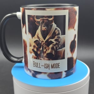 May include: A white ceramic mug with a black handle and a cow-print design. The mug features a framed image of a bull wearing a robe, with the text "BULL-ISH MODE". The mug sits on a blue and white surface.