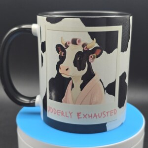 May include: A ceramic coffee mug with a black handle and rim, featuring a black and white cow print. The mug displays a cow wearing curlers and a pink robe, with the text "UDDERLY EXHAUSTED".