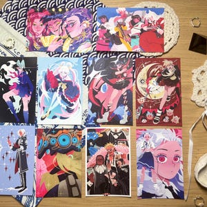May include: A collection of eleven rectangular art prints featuring diverse anime-style character illustrations in vibrant colours, including pink, blue, red, and black. The prints are arranged on a surface alongside a white fabric bag with blue wave patterns, two white crocheted coasters, a small black pyramid, and a white ceramic bowl containing a clear crystal.