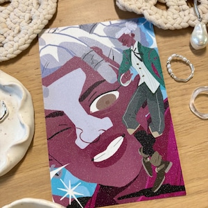 Puede incluir: Una colorida impresi&oacute;n art&iacute;stica con una ilustraci&oacute;n estilizada de una persona con chaqueta verde y chaleco blanco, sobre un gran rostro. La impresi&oacute;n tiene un aspecto brillante y texturizado, colocada sobre una superficie de madera, rodeada de joyas y objetos decorativos.