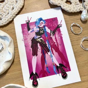 Puede incluir: Ilustraci&oacute;n vibrante de un personaje con cabello azul, sobre un fondo rosa. El personaje lleva un atuendo negro y tacones altos. La obra de arte est&aacute; en una tarjeta blanca, rodeada de objetos decorativos.