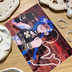 Puede incluir: Una tarjeta de arte impresa que presenta un personaje con cabello azul y una expresi&oacute;n traviesa. La obra de arte tiene un fondo rojo oscuro y marr&oacute;n con detalles de l&iacute;neas blancas. El personaje lleva un atuendo negro con detalles dorados. La tarjeta est&aacute; rodeada de joyas y objetos decorativos.