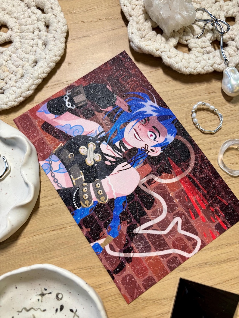 Puede incluir: Una tarjeta de arte impresa que presenta un personaje con cabello azul y una expresi&oacute;n traviesa. La obra de arte tiene un fondo rojo oscuro y marr&oacute;n con detalles de l&iacute;neas blancas. El personaje lleva un atuendo negro con detalles dorados. La tarjeta est&aacute; rodeada de joyas y objetos decorativos.