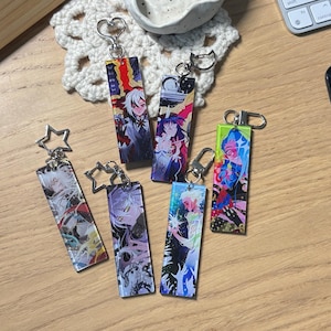 May include: Six rectangular acrylic keychains featuring colourful anime-style character illustrations. Each keychain has a metal clasp, with some shaped like stars and others like hearts. The designs include vibrant artwork with various characters.