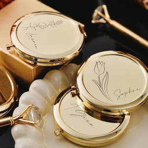 以下が含まれることがあります： 金色のコンパクトミラー。彫刻された花の模様と名前が特徴です。あるミラーには小枝、別のミラーにはチューリップ、3つ目にはシンプルな筆記体があります。ミラーは白いスカラップの皿の上に配置されています。