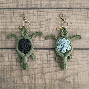 May include: Two macrame turtle keychains. One has a black center, the other a light blue center. Both are olive green with gold-colored hardware. One has a small bell attached. The keychains are on a wooden surface.