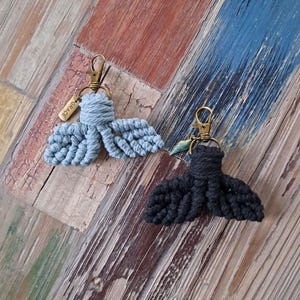 May include: Two macrame keychains, one light blue and one black, each featuring a tail design. The keychains have a gold-colored clasp and a small tag that reads "GRACE". The keychains are displayed on a wooden surface.