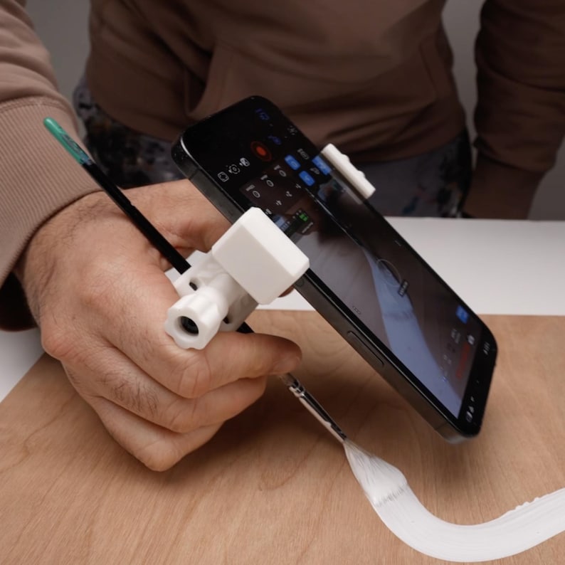 May include: A white smartphone holder with a black smartphone and a paintbrush. The holder is attached to the phone and a paintbrush, allowing for hands-free painting or drawing. The setup is on a wooden surface.