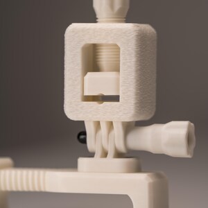 May include: Off-white camera mount with a textured surface. The mount features a rectangular body with a threaded opening, a pivoting joint, and a cylindrical extension with a ribbed knob. The design includes a screw-in top and a base with a curved edge.