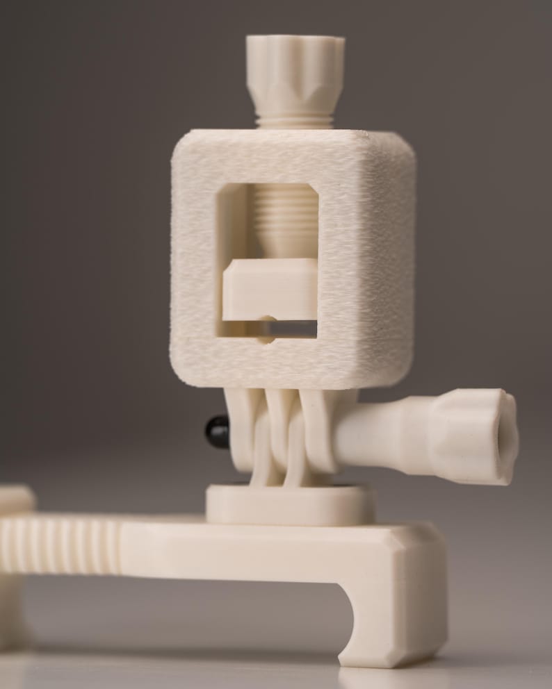 May include: Off-white camera mount with a textured surface. The mount features a rectangular body with a threaded opening, a pivoting joint, and a cylindrical extension with a ribbed knob. The design includes a screw-in top and a base with a curved edge.