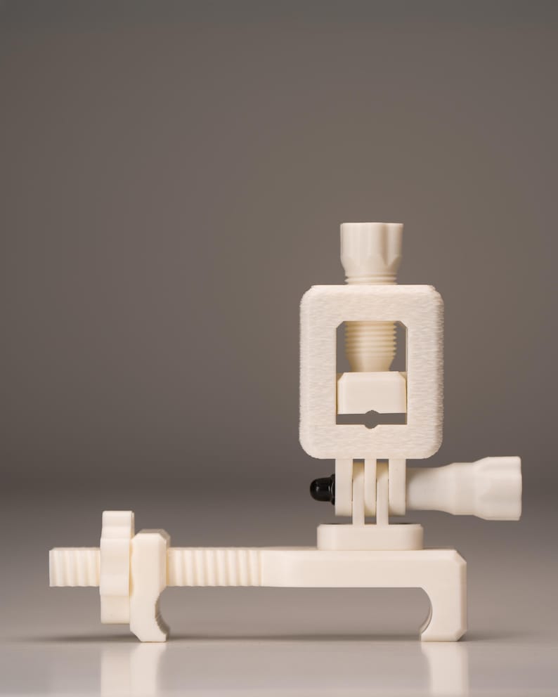 May include: A cream-colored, 3D-printed clamp with a screw mechanism. The clamp features a rectangular frame, a threaded rod, and a handle. The design is minimalist and functional, suitable for holding objects.