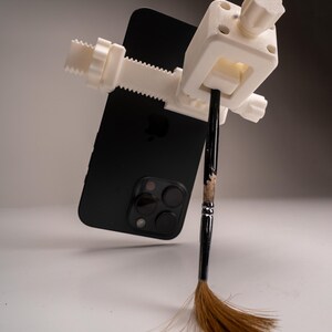 May include: A white 3D-printed clamp holding a black smartphone and a paintbrush. The clamp has a screw mechanism for adjusting the grip. The paintbrush has a black handle and brown bristles.