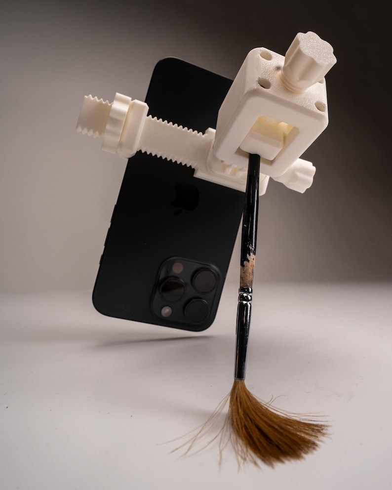 May include: A white 3D-printed clamp holding a black smartphone and a paintbrush. The clamp has a screw mechanism for adjusting the grip. The paintbrush has a black handle and brown bristles.